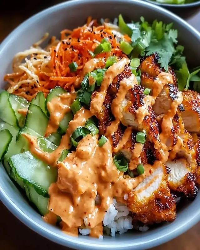 A close-up of a bang bang chicken bowl showing seasoned chicken, fresh vegetables, and creamy bang bang sauce drizzled on top